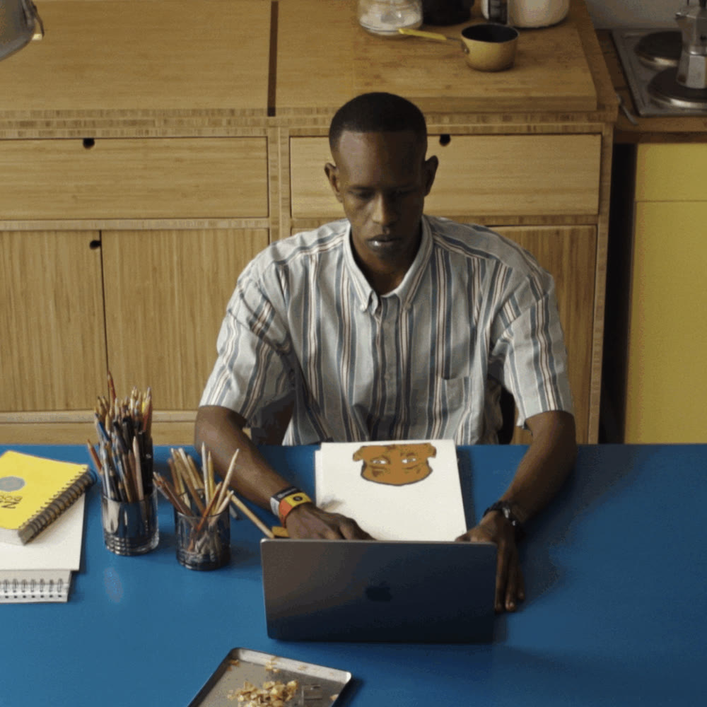 Chris-Armel Iradukunda at his kitchen table, working at a laptop with sketchbooks and pencils, a paper rendering of a face on his right